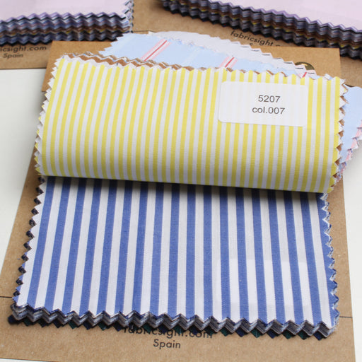 A stack of folded fabric swatches on kraft paper cards, with the top swatch rolled up. The most prominent swatch has yellow and white vertical stripes, and another underneath has blue and white vertical stripes.