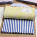 A stack of folded fabric swatches on kraft paper cards, with the top swatch rolled up. The most prominent swatch has yellow and white vertical stripes, and another underneath has blue and white vertical stripes.